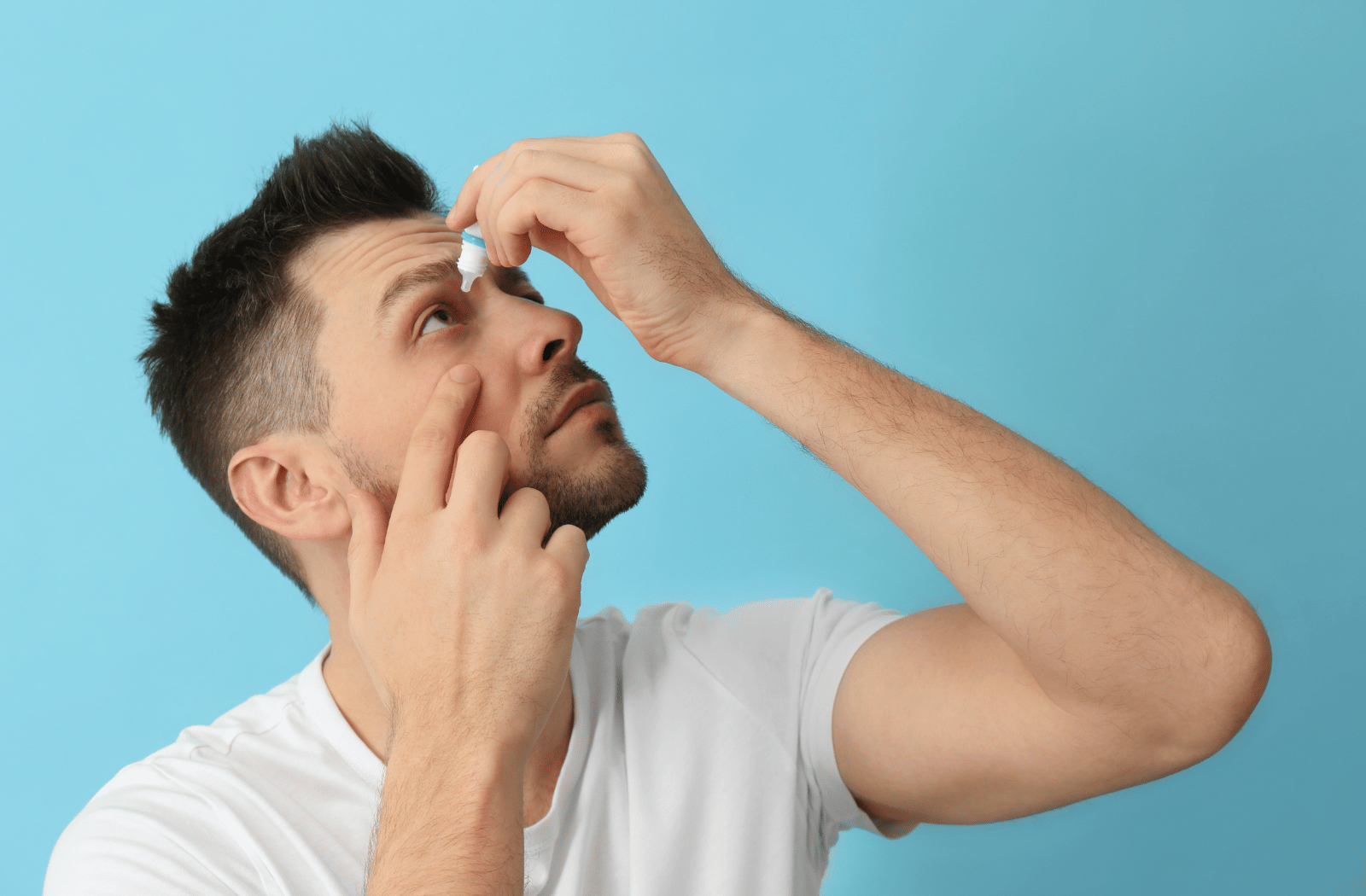 A man standing with a bottle of eye drops in his hand. He is tilting his head back and using the eye drops to treat his eyes