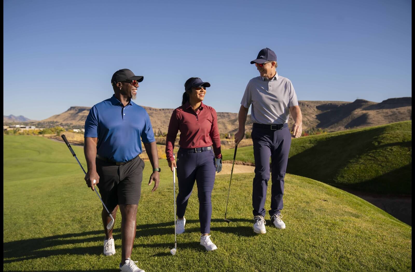 Three people, two men and one woman, walking on a sunny golf course with golf clubs in hand, wearing athletic golf attire and sunglasses, smiling and chatting with a scenic mountainous backdrop.