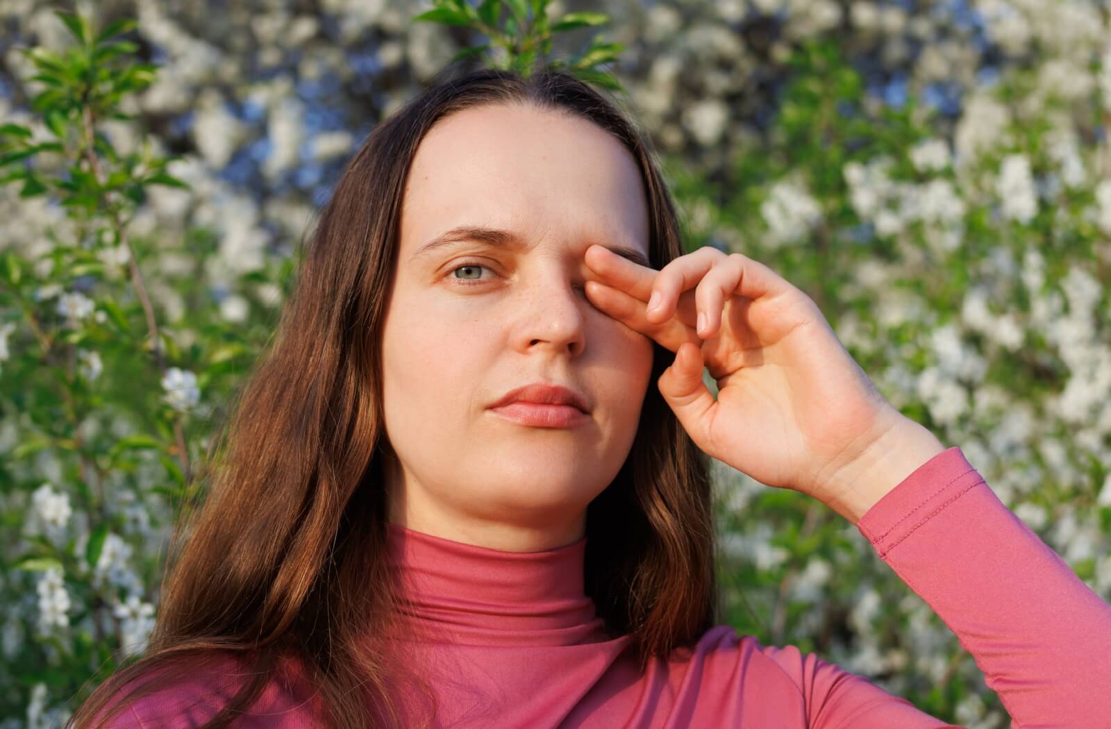 Person in pink top rubbing one eye while standing outdoors near blooming white flowers