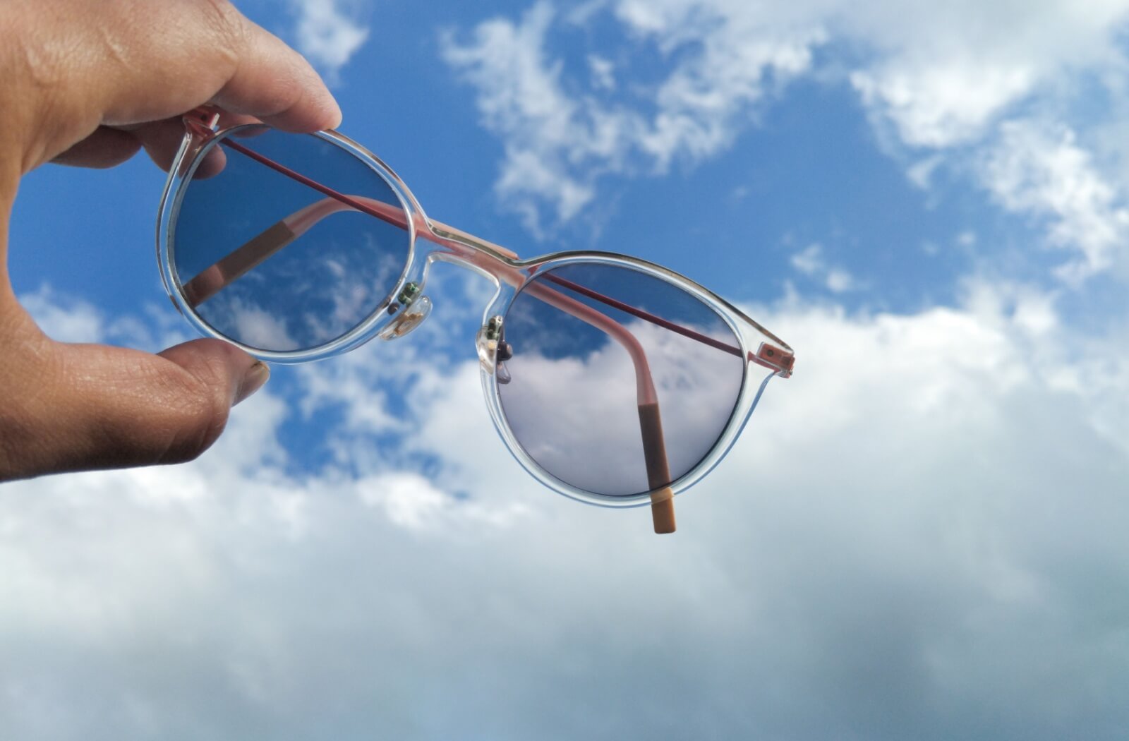 A hand holding photochromic sunglasses against a bright blue sky.