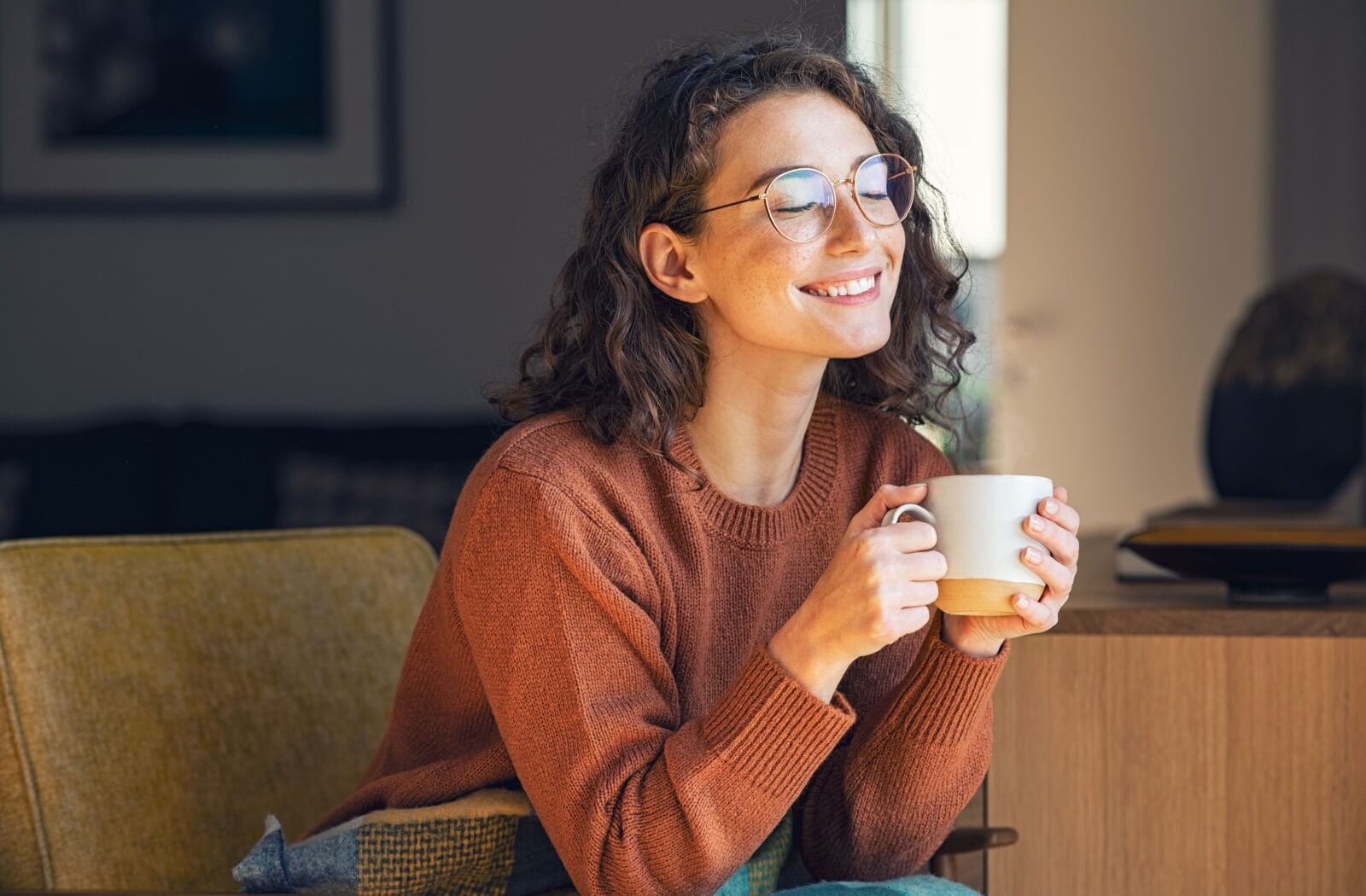 A young adult wearing a new pair of eyeglasses smiles over a cup of warm coffee in the morning.