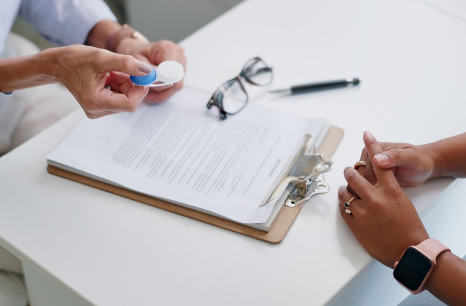 Optometrist holding a contact lens case while discussing lens care with a patient seated across a desk, with paperwork and glasses on the table.